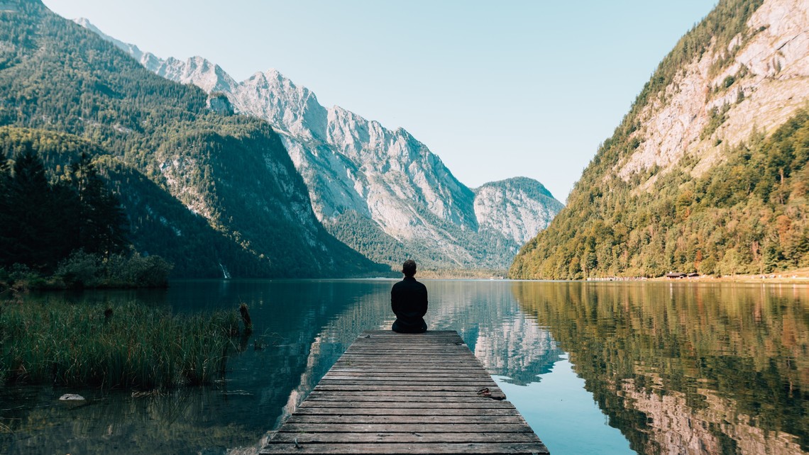 Person sitting on a dock near a lake surrounded by mountains